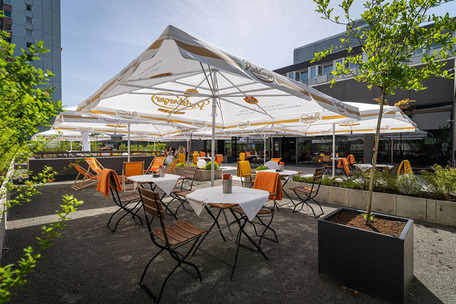 Außensitzbereich eines Hotels mit weiß gedeckten Tischen, Holzstühlen mit orangen Decken, weißen Sonnenschirmen und Pflanzen.