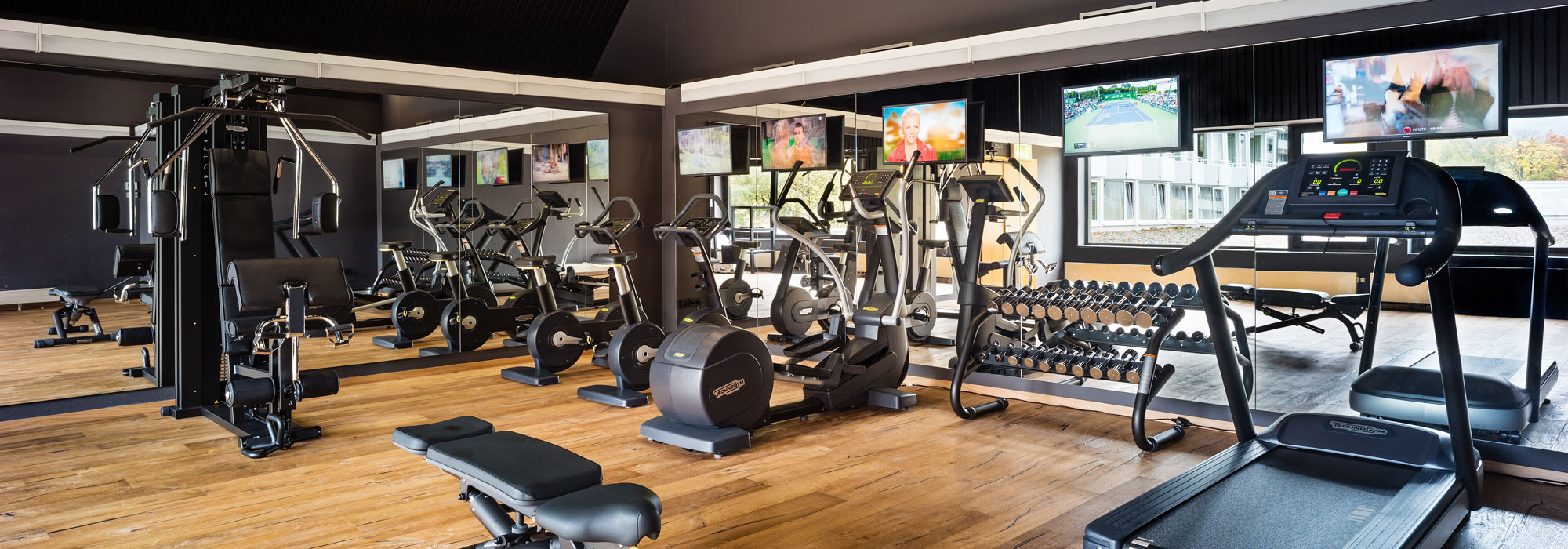 Moderner Fitnessraum mit Laufbändern, Fahrrädern, Kraftgeräten und Hanteln auf hellem Boden.