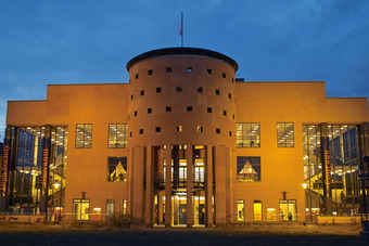 Pfalztheater Kaiserslautern bei Abendbeleuchtung mit rundem Mittelbau, Glasfassaden und Bannern, darunter einer mit „Courage“.