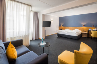 Hotelzimmer mit Doppelbett, Sofa, gelben Sesseln, Tisch mit Wasserflasche, Schreibtisch und Fernseher