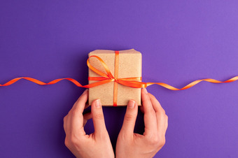 Zwei Hände halten Geschenk mit orangenem Band vor einem lila Hintergrund